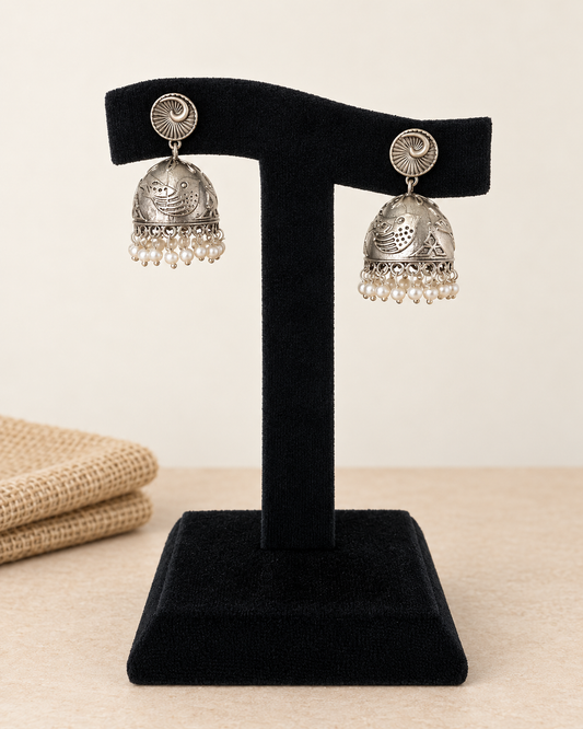 A pair of silver-toned traditional jhumka earrings with dangling pearl-like beads, displayed on a black velvet jewelry stand against a warm beige background.