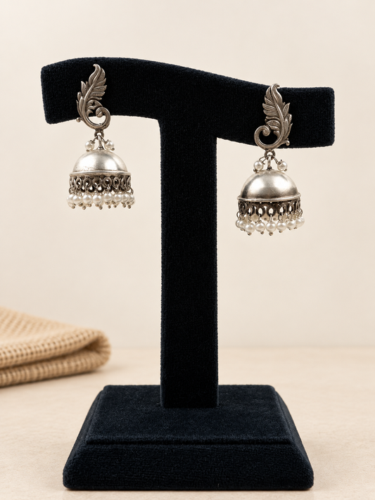 A pair of silver-toned traditional jhumka earrings displayed on a black velvet T-stand against a warm beige background.