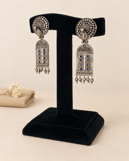 A pair of silver-toned traditional Indian jhumka earrings with blue stone accents, displayed on a black velvet jewelry stand against a warm beige background.