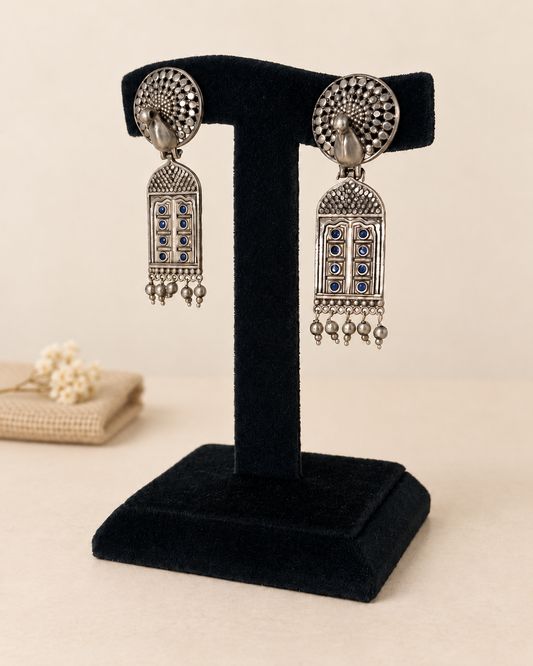 A pair of silver-toned traditional Indian jhumka earrings with blue stone accents, displayed on a black velvet jewelry stand against a warm beige background.