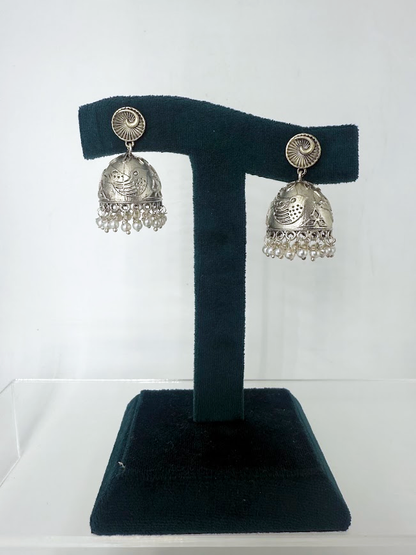 A pair of silver-toned Indian jhumka earrings with intricate filigree work and dangling bead fringes, displayed on a black velvet T-stand against a white background.