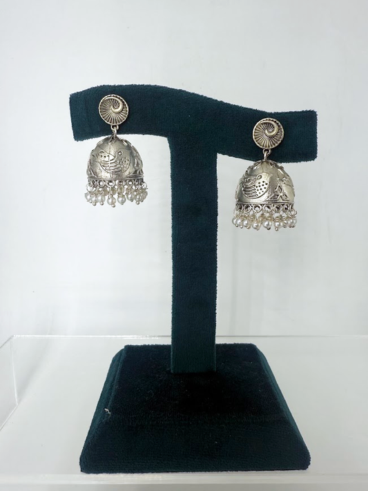 A pair of silver-toned Indian jhumka earrings with intricate filigree work and dangling bead fringes, displayed on a black velvet T-stand against a white background.