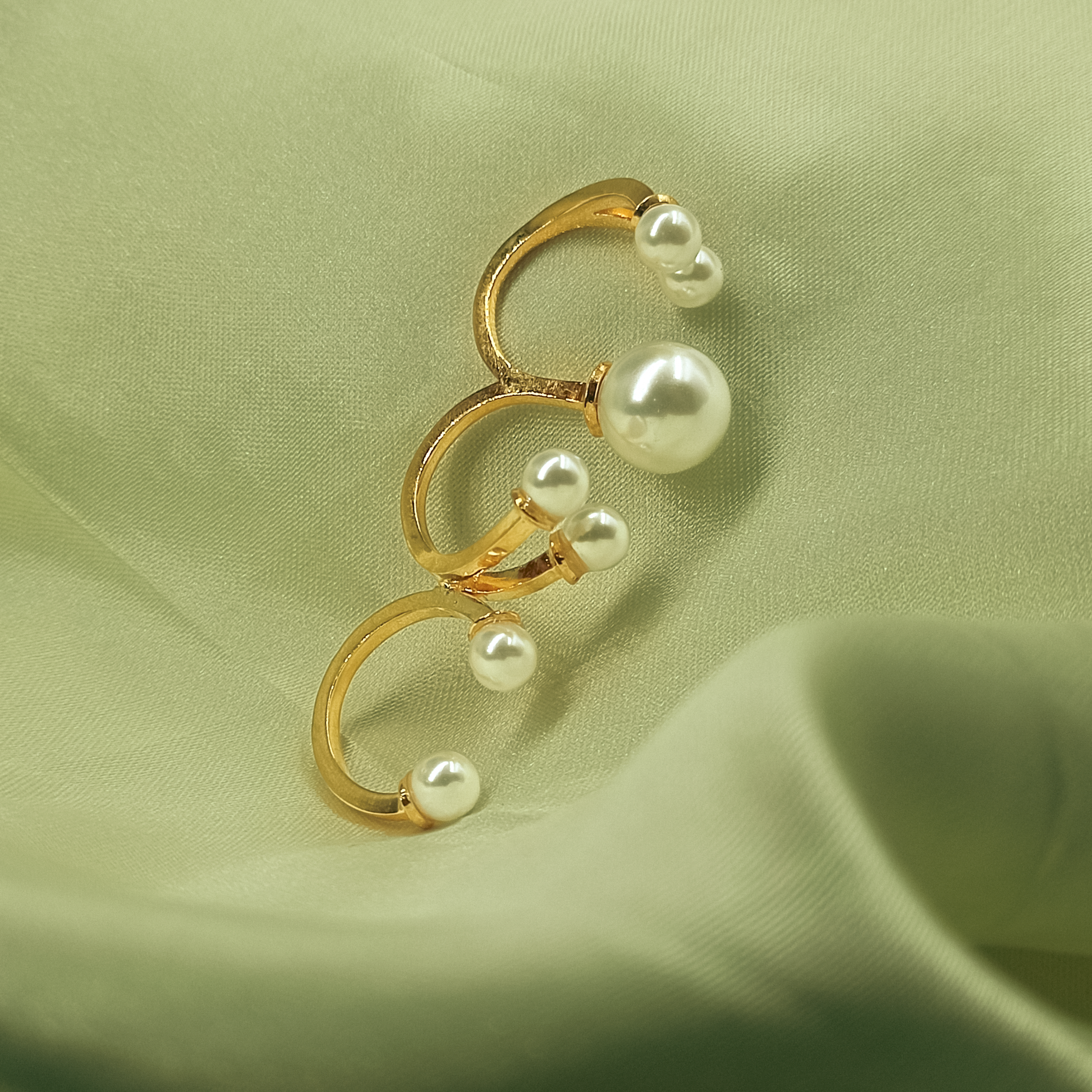 A gold-tone spiral ring earring decorated with multiple white faux pearls, displayed on a draped light sage green fabric background.