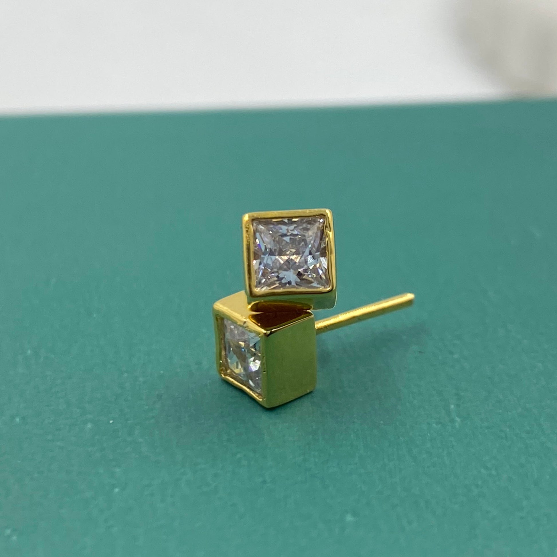 A pair of gold stud earrings with square princess-cut clear gemstones, displayed on a textured teal surface.