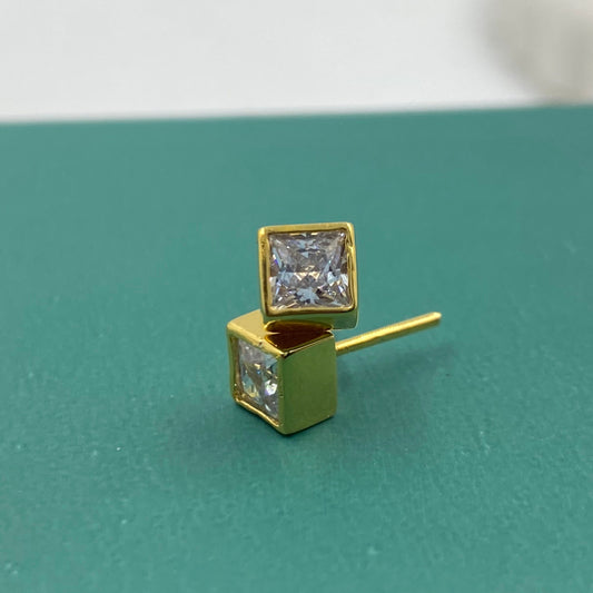 A pair of gold stud earrings with square princess-cut clear gemstones, displayed on a textured teal surface.