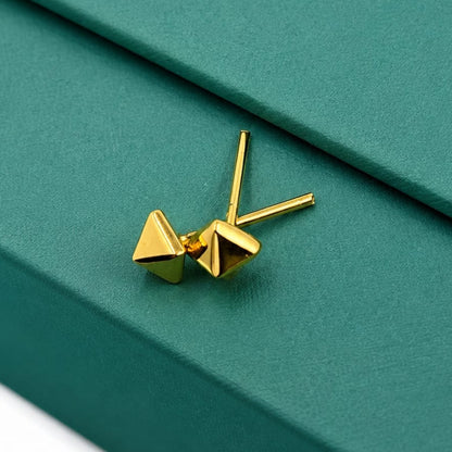 A pair of gold geometric stud earrings with triangular pyramid-shaped fronts and straight post backs, displayed on a textured dark green jewelry box.