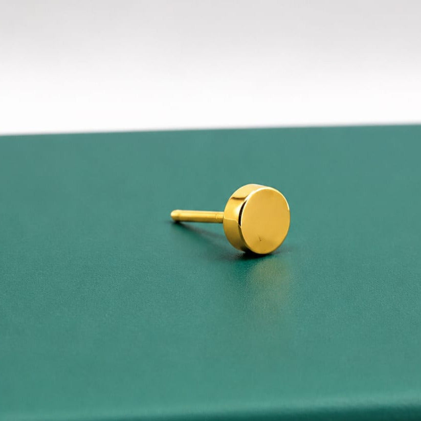 A single gold stud earring with a flat circular disc face and a straight post, resting on a dark green surface against a white background.