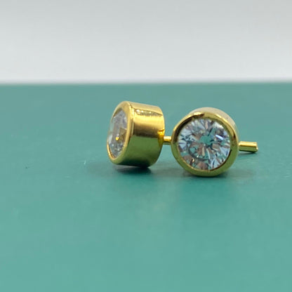A pair of gold stud earrings with round bezel-set clear gemstones, shown on a teal surface with a white background.